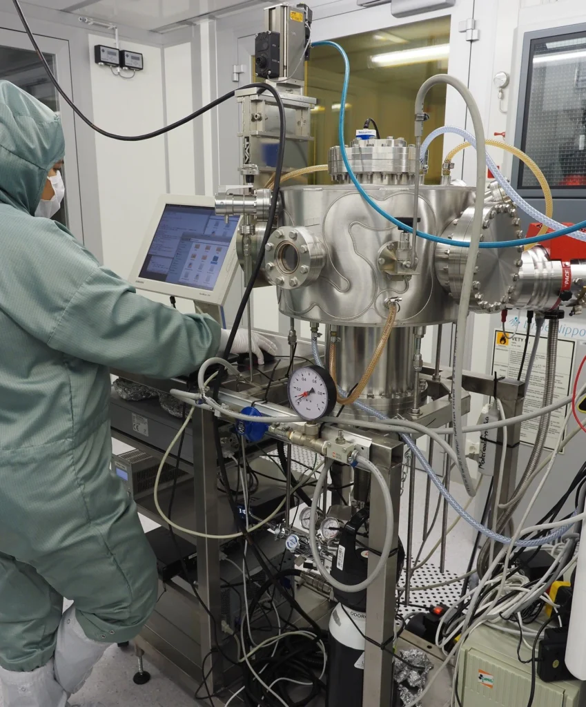 Scientist in cleanroom suit operating high-vacuum semiconductor equipment with touchscreen interface and complex tubing system